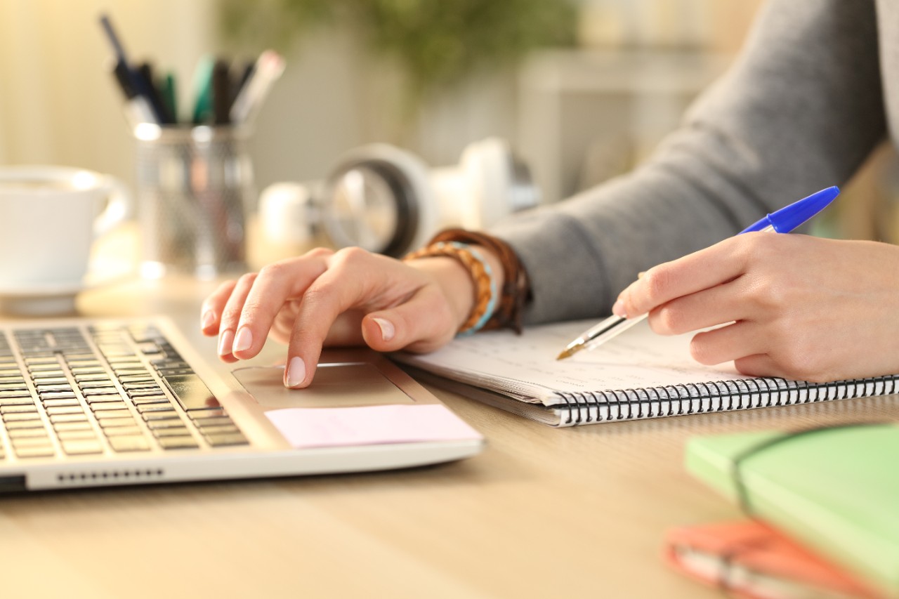 Close up of student girl hands comparing notes on notebook with laptop at home